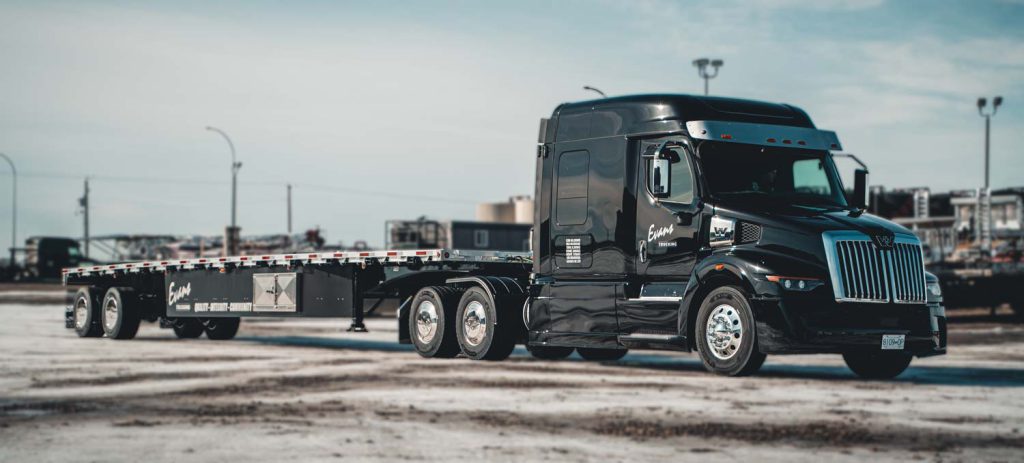 A black Evans Trucking semi truck with a flatbed trailer is parked on a lot with a lightly snow-covered ground. The truck and trailer are clean and polished, with the Evans Trucking logo visible on the side. The background includes industrial buildings and equipment.
