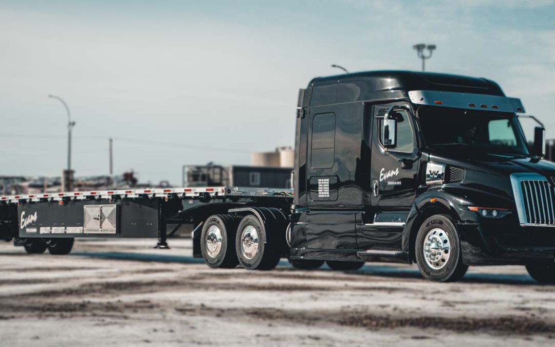 A black Evans Trucking semi truck with a flatbed trailer is parked on a lot with a lightly snow-covered ground. The truck and trailer are clean and polished, with the Evans Trucking logo visible on the side. The background includes industrial buildings and equipment.