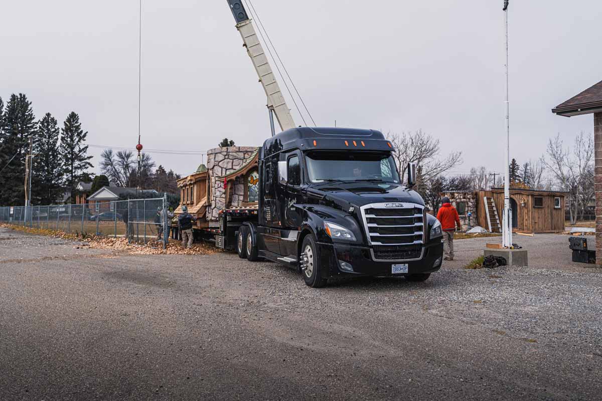 Horizontal – Charmed Delivery-3 Evans Trucking black semi with RGN trailer delivering a playhouse at a construction site with a crane and workers.